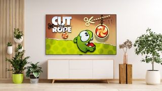 The home screen of Cut the Rope is on the TV in a light room. There are houseplants on the floor and drawers below the TV.