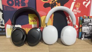 White Sonos Ace and black Bose QuietComfort Ultra Headphones standing side by side on a wooden table in front of some vinyl sleeves.